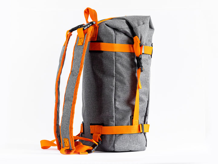 Side view of the Grava Adventure Backpack.  We can see the side stitching of the grey canvas material.  The roll top has a strap connecting the top and bottom with an easy clip system.  The two matching shoulder straps are seen from a side profile view.