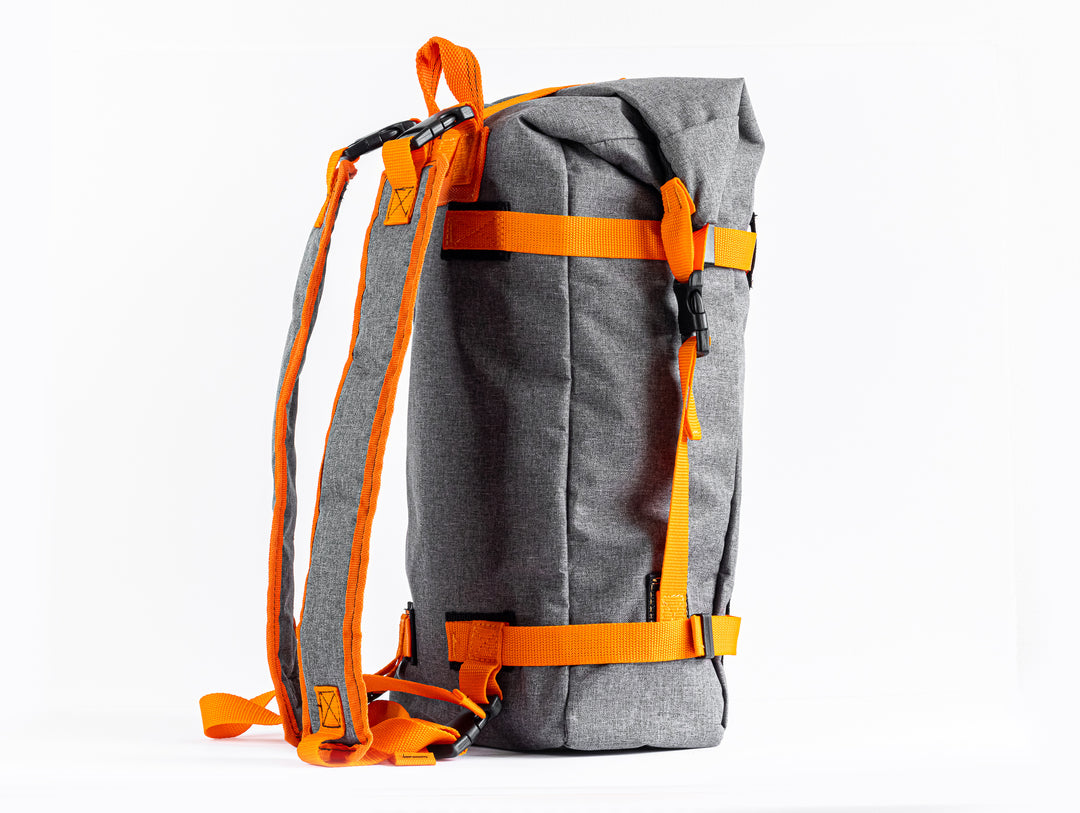 Side view of the Grava Adventure Backpack.  We can see the side stitching of the grey canvas material.  The roll top has a strap connecting the top and bottom with an easy clip system.  The two matching shoulder straps are seen from a side profile view. 