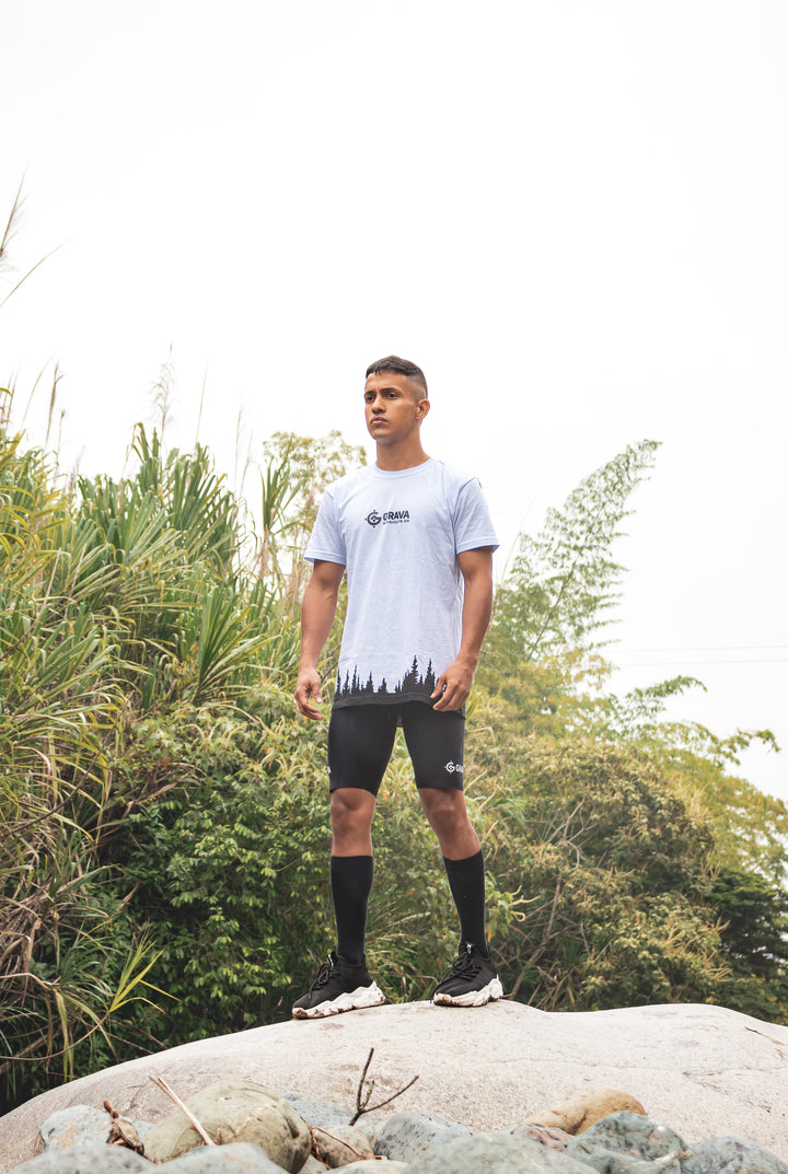 A young man wearing the Grava Mountain Shirt, standing confidently on a rock formation. He gazes into the distance, showcasing a sense of adventure and appreciation for the mountains. The shirt's design, featuring the Grava logo and a printed mountain range, resonates with his surroundings, reflecting his love for nature and exploration.