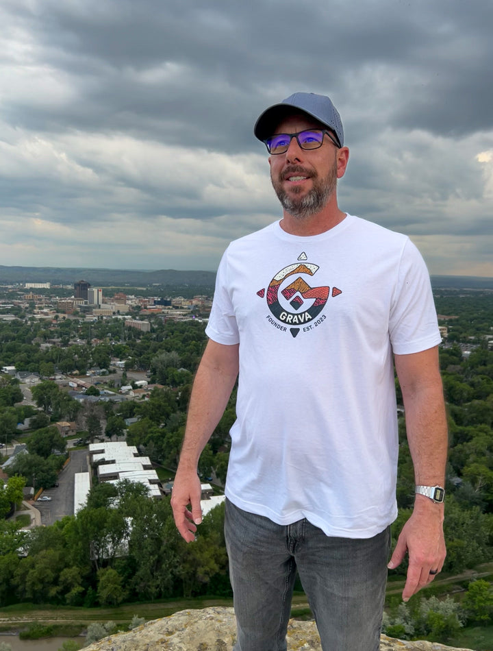 A man standing confidently on a cliff, overlooking a sprawling cityscape, while proudly wearing the Founder's T-Shirt. The vibrant colors and intricate design of the shirt's Grava Logo symbolize the spirit of adventure and exploration. 