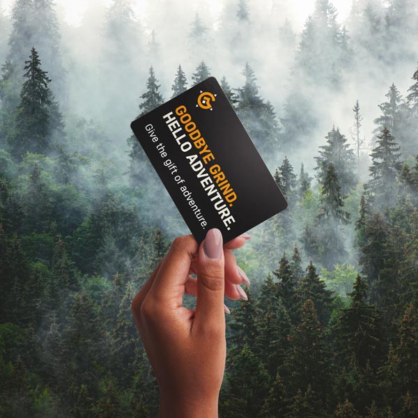 A hand holding up a Grava Adventure Gift Card against a backdrop of lush trees. The gift card is prominently displayed, featuring the Grava logo and an adventure-themed design. The hand holding the card signifies the act of giving and the excitement of receiving this special gift. The image captures the essence of the Grava Adventure Gift Card, representing the opportunity to escape the daily grind and embrace the beauty of nature. 